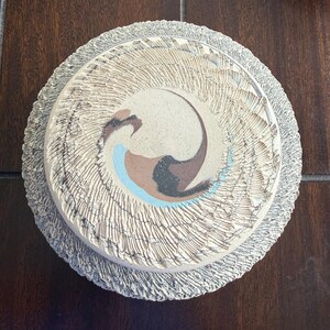 May include: A circular ceramic plate featuring a textured, carved outer rim in a light grey tone. The centre displays a smooth, flat surface with a swirling abstract design in shades of cream, brown, black, and light blue, creating a dynamic pattern.