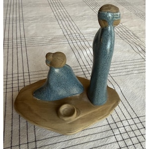 May include: A ceramic figurine set featuring two stylized figures with blue speckled robes and brown hair, positioned on a smooth, tan, oval-shaped base. A small, circular, bowl-like indentation is located on the base in front of the figures.
