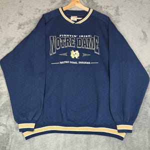 Puede incluir: Una sudadera azul marino de manga larga con el texto "FIGHTIN' IRISH", "NOTRE DAME" y "NOTRE DAME, INDIANA" en letras blancas y doradas. La prenda incluye un logotipo dorado de Notre Dame, un cuello acanalado y puños y dobladillo a rayas.