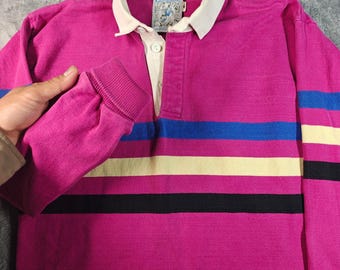 Puede incluir: Una camisa de rugby de manga larga en color magenta con cuello blanco y tapeta de botones. La prenda presenta rayas horizontales en azul, crema y negro en el pecho y las mangas. Se observa una etiqueta de Lands' End en el cuello.