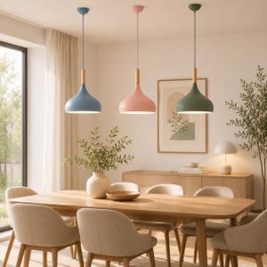 May include: Three pendant lights with bell-shaped shades in matte blue, pink, and green. Each fixture features a light-toned wooden cylindrical connector at the top, suspended by a thin cord from the ceiling above a wooden dining table and chairs.