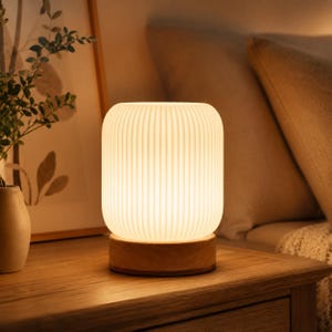 Puede incluir: Una lámpara de mesa cilíndrica con una pantalla blanca translúcida de costillas verticales que emite un brillo cálido. La lámpara se apoya sobre una base circular de madera clara, proporcionando una base estable para el objeto iluminado.