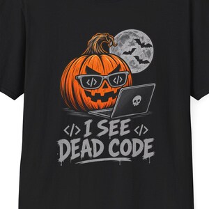May include: A black short-sleeved t-shirt featuring a graphic of an orange jack-o'-lantern wearing glasses with code brackets, sitting at a laptop with a skull logo. Above is a full moon with bats. The text reads I SEE DEAD CODE.