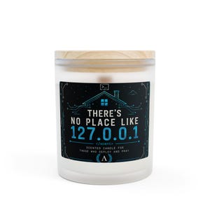 Könnte beinhalten: Eine Kerze im Milchglas mit einem hellen Holzdeckel. Das schwarze Etikett zeigt eine blaue Hausgrafik aus Leiterbahnen und den Text: THERE'S NO PLACE LIKE 127.0.0.1 </scent> SCENTED CANDLE FOR THOSE WHO DEPLOY AND PRAY. Ein Terminal-Symbol ist oben.