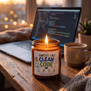 Op de afbeelding: Een brandende kaars in een amberkleurige glazen pot met een wit etiket met de tekst "SMELLS LIKE CLEAN CODE" in blauwe en groene letters, omringd door kleine iconen en de tekst "CODE WITH CODE" en "GOOD CODE GOOD VIBES".