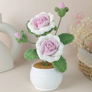 May include: A crochet flower arrangement featuring two blooming roses with light pink centers and white outer petals, accompanied by two small pink buds and green leaves, all set in a round white crocheted pot with a tan top surface.