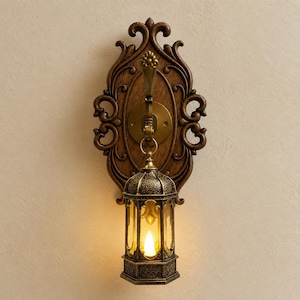 Op de afbeelding: Een wandlamp met een donkerbruine, sierlijke houten achterplaat met krulwerkdetails. Een messingkleurige metalen haak steekt uit het midden en ondersteunt een hangende, zeshoekige lantaarn met een warm gloeiende led-kaars erin.