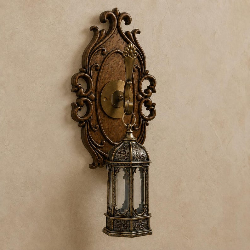 Op de afbeelding: Een wandlamp met een donkerbruine, sierlijke houten achterplaat met krulwerk. Een messingkleurige metalen haak steekt uit het midden en draagt een kleine, zeshoekige lantaarn met ingewikkelde metalen filigraanpatronen.