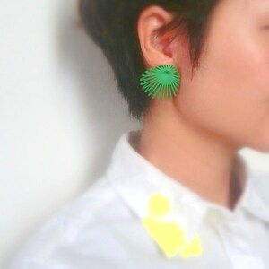 May include: A pair of green geometric earrings with a fan-like design. The earrings are worn by a person with short dark hair.
