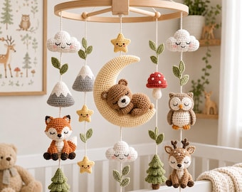 May include: A crochet baby mobile featuring a wooden circular frame with hanging plush figures including a brown bear sleeping on a yellow crescent moon, a fox, an owl, a deer, white clouds, green pine trees, a red mushroom, and yellow stars.