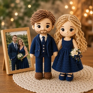 Puede incluir: Dos muñecos de ganchillo que representan a una pareja. El muñeco masculino viste un traje azul marino con camisa blanca y zapatos marrones. La muñeca femenina viste un vestido azul marino y sostiene un pequeño ramo de flores blancas y azules.