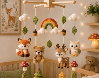 May include: A hanging mobile featuring crocheted figures of a fox, bear, deer, and owl, accented with a rainbow, clouds, acorns, pine trees, and leaves. The mobile is suspended from a light-colored wooden frame with white strings.
