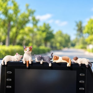 May include: Five small cat figurines perched on the top edge of a black vehicle dashboard screen. The cats include a white sleeping kitten, a sitting calico, a gray and white cat, a brown and white cat, and a brown and white sleeping kitten.