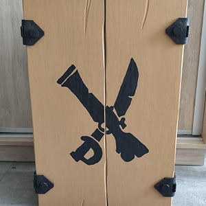 May include: A rectangular wooden cabinet with a light tan finish and vertical wood grain texture. The front features a black graphic of a crossed sword and flintlock pistol. Four black metal corner brackets with visible bolts secure the edges.
