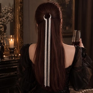 Könnte beinhalten: Ein silberfarbenes Haaraccessoire mit einer halbmondförmigen Spange und zwei langen, herabhängenden Metallkettchen, die vertikal in der Mitte des Haares verlaufen. Das Accessoire besteht aus polierten Metallgliedern.