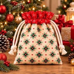 May include: A fabric drawstring gift bag featuring a cream-colored base with a repeating pattern of green and red snowflakes and small red hearts. The top ruffle is red with white snowflakes, and the bag includes thick white cotton rope ties.