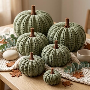 May include: A collection of six sage green crocheted pumpkins of varying sizes, each featuring a textured ribbed pattern and a solid brown stem. The pumpkins are arranged on a cream-colored knitted runner alongside small decorative leaves.