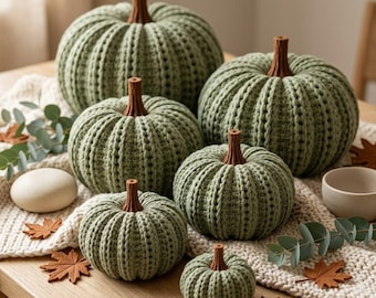 May include: A collection of six sage green crocheted pumpkins of varying sizes, each featuring a textured ribbed pattern and a solid brown stem. The pumpkins are arranged on a cream-colored knitted runner alongside small decorative leaves.
