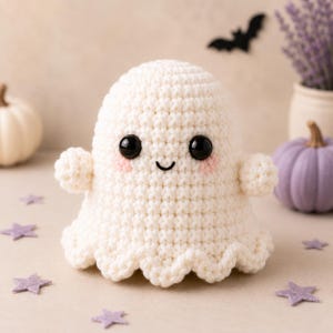 May include: A cream-coloured crocheted ghost plushie featuring a rounded head, two small arms, and a scalloped bottom edge. The ghost has large, glossy black safety eyes, a small embroidered black mouth, and soft pink blush on its cheeks.