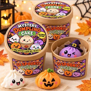 May include: Two round cardboard containers labelled Mystery Dumpling Cake Halloween Edition with cartoon graphics. One open container reveals a purple dumpling wearing a witch hat. Two additional dumplings, one white and one orange, sit in front.