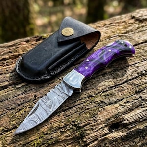 Könnte beinhalten: Ein Klappmesser mit einer Damaststahlklinge mit Wellenmuster, einer polierten silbernen Backe und einem violett marmorierten Griff mit drei silbernen Stiften. Neben dem Messer liegt eine schwarze Lederscheide mit einem goldfarbenen Druckknopf.
