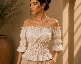 May include: A beige off-the-shoulder blouse featuring a ruffled neckline, smocked waist, and elbow-length sleeves with ruffled cuffs. The garment is crafted with a gathered bodice and a flared hem, providing a structured yet fluid silhouette.