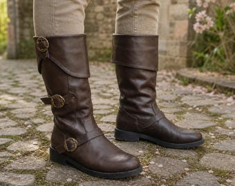May include: A pair of dark brown mid-calf boots featuring a folded cuff at the top. The boots are adorned with three decorative circular buckles with an intricate textured pattern, positioned along the side and ankle area of the footwear.