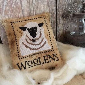 May include: A white sheep with black eyes and a brown nose is embroidered on a tan pillow with the word "Woolens" stitched in black. The pillow is sitting on a white fluffy surface.