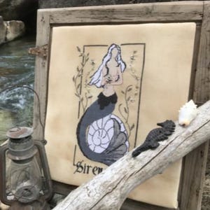 Könnte beinhalten: Ein gerahmtes Stickbild auf cremefarbenem Stoff zeigt eine Meerjungfrau mit langem weißem Haar und dunklem Schwanz innerhalb eines rechteckigen Rahmens mit dem Wort Siren darunter. Eine Metalllaterne und Treibholz befinden sich daneben.