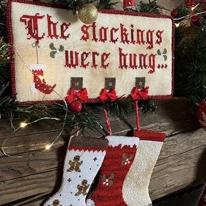 Op de afbeelding: Een witte kruissteek sampler met de tekst "The stockings were hung..." en vier rood-witte kousen die eronder hangen. De kousen zijn versierd met peperkoekmannetjes, hulst en sneeuwvlokken.