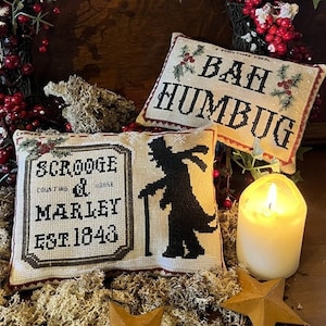 Puede incluir: Tres fundas de cojín bordadas con un tema de villancico navideño. La primera funda de cojín presenta una silueta de Ebenezer Scrooge, la segunda funda de cojín dice "Scrooge & Marley Est. 1843", y la tercera funda de cojín dice "Bah Humbug".