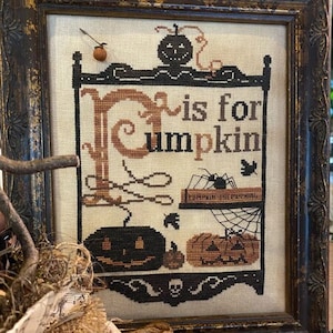 Op de afbeelding: Een ingelijst kruissteekontwerp met de tekst "P is for Pumpkin" en een zwarte en oranje pompoen met een jack-o'-lantern gezicht. De lijst is van donkerbruin hout met een verweerde afwerking. Halloween-collectie 2021 van The Primitive Hare.