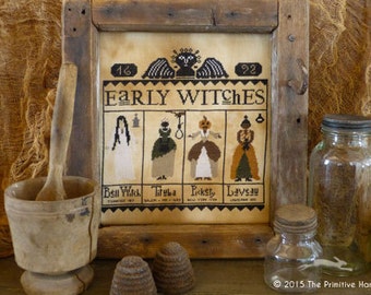 May include: A framed cross-stitch sampler with the title "Early Witches" and four witches depicted in individual squares. The witches are named Bell Witch, Tituba, Pickety, and Laveau. The sampler is displayed on a wooden surface with other decorative items.