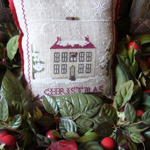 Peut inclure: Un oreiller blanc avec une bordure rouge présente un motif de point de croix d'une maison avec de la neige sur le toit et le mot "NOËL" en bas. L'oreiller est entouré de feuilles vertes et de baies rouges.