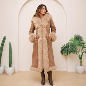 May include: A long, tan suede coat featuring a thick, shaggy beige faux fur trim along the collar, front opening, cuffs, and hem. The coat has a fitted silhouette with long sleeves and two visible side pockets, paired with dark brown leather boots.