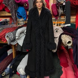 May include: A long black coat featuring a thick, textured black fur-like trim along the wide lapels, the sleeve cuffs, and the bottom hem. The coat has a smooth, matte finish and reaches down to the lower calf, paired with black pointed-toe shoes.