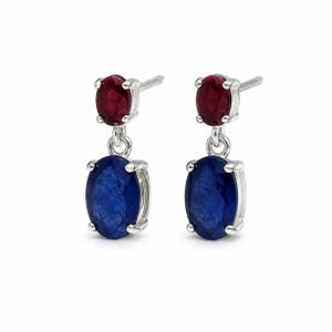Könnte beinhalten: Ein Paar Ohrhänger mit einem kleinen oval geschliffenen roten Edelstein über einem größeren oval geschliffenen blauen Edelstein. Jeder Stein ist in einer silberfarbenen Metallfassung mit vier Krappen gefasst und besitzt einen Steckerverschluss.