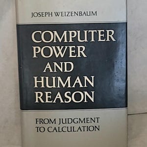 Może przedstawiać: Książka w twardej oprawie zatytułowana Computer Power and Human Reason: From Judgment to Calculation autorstwa Josepha Weizenbauma. Okładka posiada ciemnoniebieski panel środkowy z białym tekstem, otoczony jasnoszarymi sekcjami na górze i na dole.