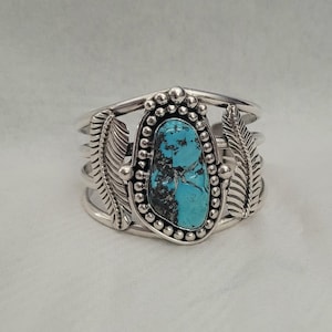 May include: A silver cuff bracelet featuring a central, irregular, bright blue turquoise stone with dark matrix veins. The stone is surrounded by a beaded silver border and flanked by two detailed, engraved silver feather motifs on each side.