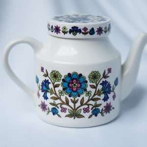 May include: A white ceramic teapot with a matching lid, featuring a colourful floral pattern in shades of blue, green, pink, and brown. The design includes a central large flower surrounded by smaller blossoms and leaves on the body and lid.