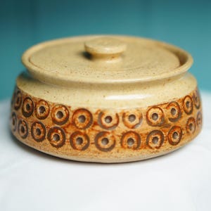 May include: A round, beige ceramic lidded jar with a speckled finish. The exterior features two horizontal rows of dark brown, circular stamped patterns encircling the base. The lid has a small, centered knob handle for easy removal.