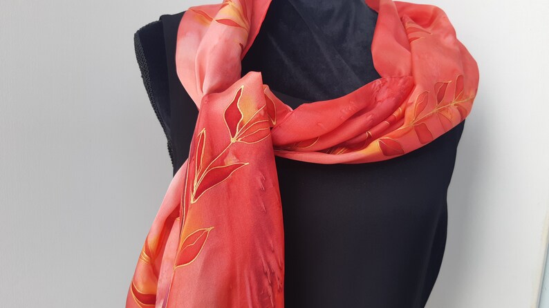 May include: A red silk scarf with a floral pattern in shades of orange and gold. The scarf is tied around a black shirt.