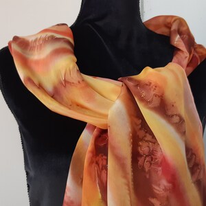 May include: A long, flowing scarf with a hand-painted design in shades of orange, yellow, and brown. The scarf is draped over a black mannequin.