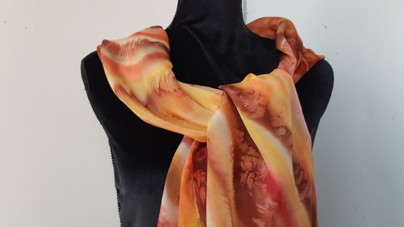 May include: A long, flowing scarf with a hand-painted design in shades of orange, yellow, and brown. The scarf is draped over a black mannequin.