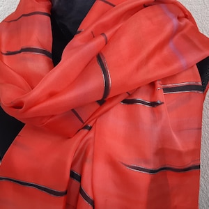 May include: A vibrant coral-colored scarf with black and silver horizontal stripes. The scarf is draped over a black form, showcasing its flowing fabric and design. This accessory adds a pop of color and pattern.