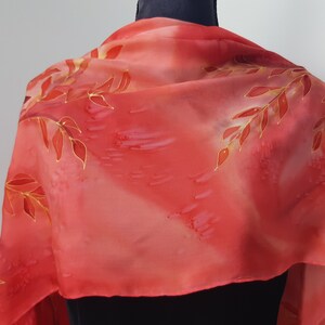 May include: A red silk scarf with a hand-painted design of gold and red leaves. The scarf is draped over a mannequin's neck.