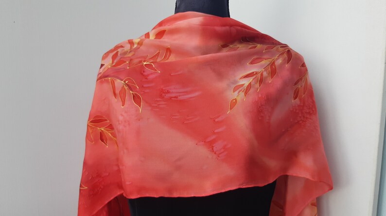 May include: A red silk scarf with a hand-painted design of gold and red leaves. The scarf is draped over a mannequin's neck.