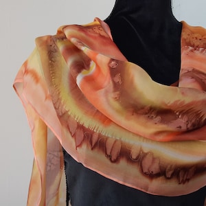 May include: A silk scarf with a hand-painted design in shades of orange, yellow, and brown. The scarf is draped over a black mannequin.