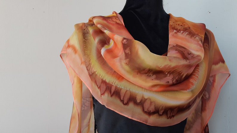 May include: A silk scarf with a hand-painted design in shades of orange, yellow, and brown. The scarf is draped over a black mannequin.