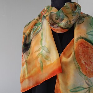 May include: A silk scarf with a hand-painted design of orange and green fruits and leaves. The scarf is a light orange color with a gradient of orange and red at the bottom.